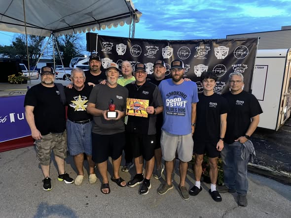 The Louie's BBQ crew at dusk under the Praise the Lard banner, holding a glass-apple trophy and the golden-ticket card